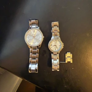 Två silverfärgade klockor - Säljer två stilrena klockor i silverfärg. Den ena har en större urtavla med romerska siffror och tre små urtavlor, medan den andra är mindre och mer diskret. Båda har länkar i metall och en klassisk design som passar till många tillfällen.