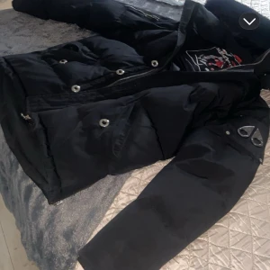 Svart dunjacka från Moose Knuckles - Säljer en svart dunjacka från Moose Knuckles med broderad logga på insidan. Jackan har dragkedja och knappar framtill samt långa ärmar. Perfekt för kyliga dagar. Storlek M möts endast upp 