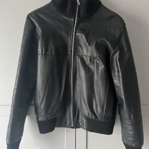 Svart skinnjacka från Paul & Friends - Snygg svart skinnjacka från Paul & Shark med dragkedja och ribbade detaljer vid krage, ärmslut och nederkant. Perfekt för en stilren look. Jackan har långa ärmar och en klassisk bomberjacka-stil.