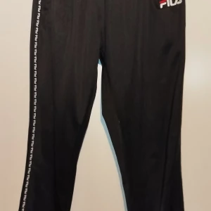 Svarta träningsbyxor från Fila - Snygga svarta träningsbyxor från Fila med logotyp längs sidorna. Tillverkade i 100% polyester för en bekväm passform. Perfekta för träning eller en avslappnad dag.