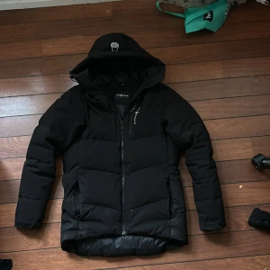 Svart pufferjacka från Sail Racing - Snygg svart pufferjacka från Sail Racing i storlek XS men passar som S. Jackan har en dragkedja framtill och en justerbar huva. Perfekt för kyliga dagar med sin vadderade design som håller dig varm. En stilren och funktionell jacka för alla tillfällen.