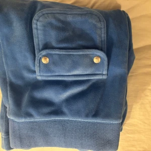 Blå hoodie från Juicy Couture - Säljer ett snyggt blått set  från Juicy Couture med dragkedja och snörning. De har en praktisk ficka med knappar på baksidan av byxa. och en broderad logga på kofta . Perfekt för en avslappnad stil.       Koftan är storlek S och byxan är i M