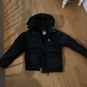 Svart dunjacka från Fjällräven - Säljer en svart dunjacka från Fjällräven Expedition Series. Jackan har en pufferdesign med långa ärmar och praktiska fickor. Perfekt för kalla dagar med sin varma och skyddande stil. Den har en dragkedja och en justerbar huva. Köpte den för 6990kr använd 1-2 gånger 
