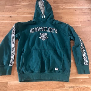 Grön Harry Potter Hogwarts hoodie 170 storlek  - Säljer en grön hoodie med Harry potter -tryck och emblem på bröstet. Den har en stor ficka fram och text längs ärmarna. Perfekt för alla Harry Potter-fans! 🧙‍♂️Som ny och jätte finnt grön .