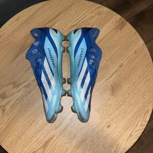 Adidas x crazy fast blåa storlek 41 1/3 41-42 - Säljer ett par blå och vita Adidas fotbollsskor med snörning och dobbar. Skorna har en sportig design med vita ränder på sidorna och är perfekta för fotbollsträning.