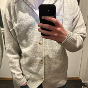 Grå kofta med huva - Säljer en skön grå kofta med huva och knappar framtill. Perfekt för kyliga dagar. Koftan har långa ärmar och ribbade muddar vid ärmslut och nederkant.