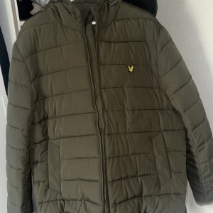 Olivgrön pufferjacka från Lyle & Scott - Säljer en olivgrön pufferjacka från Lyle & Scott med dragkedja och huva. Jackan har ett broderat emblem på bröstet och är perfekt för kyligare dagar. Den är långärmad och quiltad för extra värme. Passar bra för både höst och vinter. Har använts 2 gånger bara