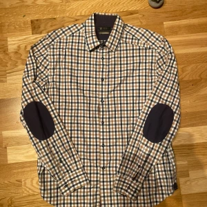 Rutig skjorta från Riley - Snygg rutig skjorta från Riley i guldigt, svart och vitt. Skjortan har långa ärmar och stiliga armbågslappar i mörkare ton. Perfekt för en avslappnad men ändå uppklädd look.