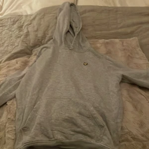 Grå hoodie från Lyle & Scott - Säljer en stilren grå hoodie från Lyle & Scott Junior. Den har en klassisk design med en liten logga på bröstet och en bekväm huva. Perfekt för en avslappnad look.
