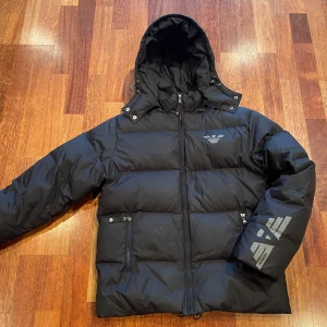 Armani jacka OG - Säljer en snygg svart dunjacka från Armani Teen. Jackan har en huva och dragkedja framtill. Den är dekorerad med det ikoniska Armani-logotypen på bröstet och ärmen. Perfekt för kalla dagar med sin vadderade design.    BUDA 