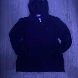 Mörkblå hoodie från GANT - Säljer en stilren mörkblå hoodie från GANT med dragkedja och broderad logotyp på bröstet. Perfekt för en avslappnad look. Passar för ungdomar runt 15 år.