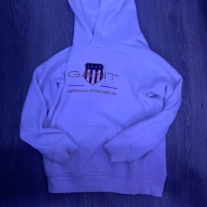  St 158-164 Vit hoodie från GANT - Säljer en stilren vit hoodie från GANT med deras klassiska logga och texten 'American Sportswear' på framsidan. Tröjan har en stor ficka framtill och en bekväm huva. Perfekt för en avslappnad stil.