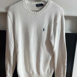 Vit stickad tröja från Ralph Lauren - Säljer en klassisk vit stickad tröja från Ralph Lauren med det ikoniska blå logotypbroderiet på bröstet. Tröjan har en rund halsringning och långa ärmar, perfekt för en stilren look. Passar bra till både jeans och chinos. Ett hål under ena armen men syns inte vid användning samt att taggen sitter något löst.