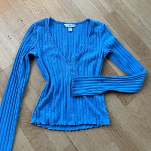 Blå ribbad topp från H&M - Säljer en snygg blå ribbad topp från H&M med v-ringning och knappar framtill. Endast använd en gång sen tvättad så toppen skick 💙extra knapp finns kvar. 63 % bomull, resten polyester & elastan