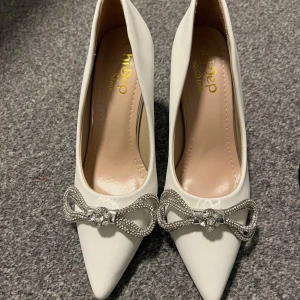Eleganta pumps med strassrosett - Snygga pumps från Chicop i både svart och vit färg. Skorna har en spetsig tå och är dekorerade med en glittrig strassrosett framtill. Perfekta för att ge en elegant touch till din outfit. Helt nya klackar aldrig använt i storlek 37.