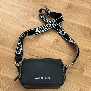 Mario Valentino väska  - Detta är en fin och mycket användbar väska med tre fack, yttre, mellan och ett litet innerfack. Väskan har lite men inte synliga skador då den har varit lite använd. Väskan är fortfarande väldigt snygg med ett justerbart band.   