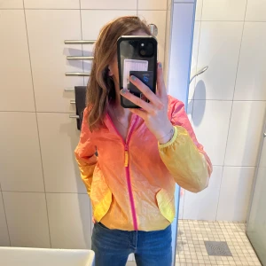 Vindjacka  - Färgglad vindjacka med fickor där fram och en stor ficka där back även till för att kunna knyckla ihop jackan till en boll. 