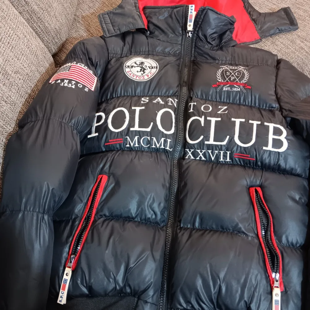 Säljer en svart pufferjacka från Santoz Polo Club med coola tryck och broderier. Jackan har en dragkedja och röda detaljer på fickorna. Insidan är röd med ett stort emblem. Perfekt för vintern och hösten!. Jackor.