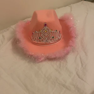 Rosa cowboyhatt med tiara - Supercool rosa cowboyhatt med en glittrig tiara framtill. Hatten har fluffiga rosa fjädrar runt brättet som ger en extra festlig touch. Perfekt för en temafest eller bara för att sticka ut i mängden. Den är i nyskick och redo för nya äventyr! bandet är justerbart!
