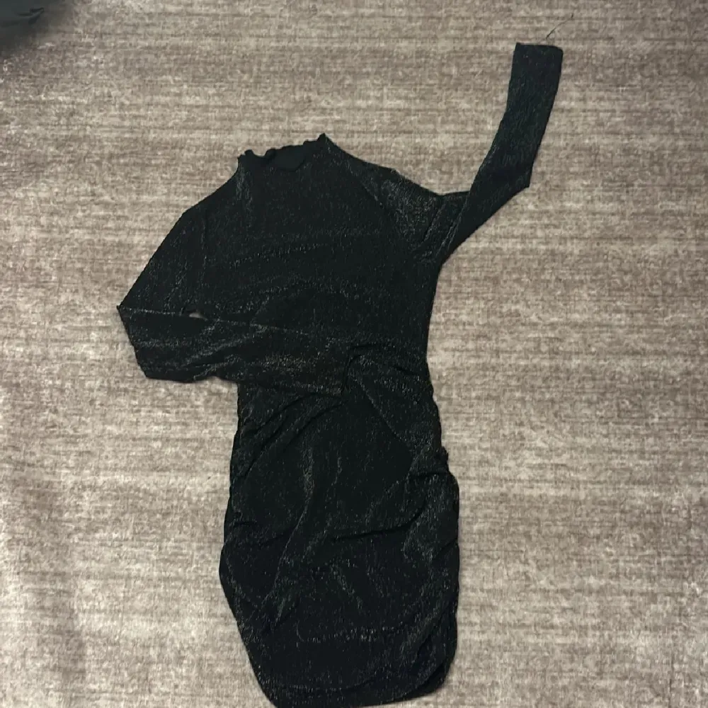 Snygg svart glittrig klänning med lång ärm och tight passform. Perfekt för en kväll ute med vänner. Klänningen har en hög hals och är kort i modellen.. Mekot.