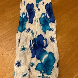 Blommig kjol med blå och vita nyanser - Säljer en somrig kjol med ett vackert blommönster i blå och vita nyanser. Kjolen har en smockad överdel och är ärmlös, perfekt för varma dagar. Den har en luftig och bekväm passform. Fungerar även som en miniklänning. Köpt från en marknad.💕🥰