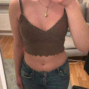 Brun ribbad linnetopp med spetsdetaljer - Säljer en brun ribbad linnetopp med spetsdetaljer längs kanterna. Toppen är croppad och har smala axelband. Perfekt för en stilren och bekväm look.