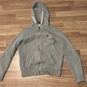Grå huvtröja från Ralph Lauren - Säljer en stilren grå huvtröja från Ralph Lauren med dragkedja och broderad logga på bröstet. Tröjan har långa ärmar och en praktisk huva med snören. Perfekt för en avslappnad look. Säljer för att den har blivit för liten för mig 