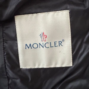 Svart dunjacka från Moncler - Säljer en snygg svart dunjacka från Moncler. Jackan har en stilren design med knappar framtill och Monclers logga på ärmen. Perfekt för kyligare dagar med sin lätta och värmande dunstoppning. Serienummer för äkthet se lilla lappen med start siffra 8….
