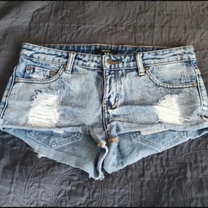 Blå jeansshorts med slitningar - Snygga blå jeansshorts från Forever 21 med coola slitningar fram och bak. De har en klassisk femficksdesign och är perfekta för en avslappnad stil. Shortsen är lågmidjade och har en rå kant vid benen för en trendig look.