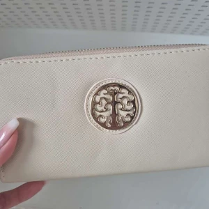 Beige plånbok med gulddetalj - Elegant beige plånbok med en stilren gulddetalj på framsidan. Plånboken har en praktisk dragkedja som håller dina saker säkra. Perfekt för att hålla ordning på kort och kontanter.