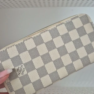 Rutig plånbok  - Säljer en stilren plånbok i ett klassiskt rutigt mönster. Plånboken är i beige och grå toner med dragkedja upptill. Perfekt för att hålla ordning på kort och kontanter.