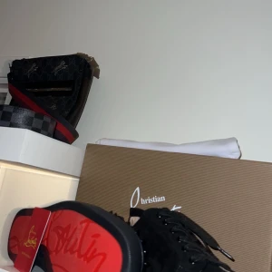 Svarta skor  från Christian Louboutin - Snygga svarta sneakers från Christian Louboutin med ikoniska röda sulor och nitar på ovansidan. Skorna har snörning och är tillverkade i mocka. Perfekta för att ge en edgy touch till din stil.