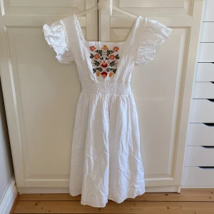 Vit klänning med blommigt broderi - Charmig vit klänning med vackert blommigt broderi i rosa, rött och gult på bröstet. Klänningen har korta puffärmar och en smockad midja för en bekväm passform. Storlek M. Använd 1-2 gånger, i fint skick. Från Shein. 