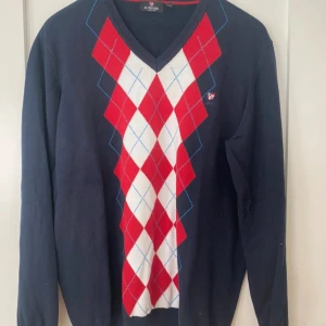 Tröja från U.S. Polo Assn. - Säljer en stilren mörkblå tröja från U.S. Polo Assn. med ett klassiskt argyle-mönster i rött och vitt på framsidan. Tröjan har en v-ringad hals och långa ärmar, perfekt för en elegant look. Passar bra till både vardag och mer uppklädda tillfällen.