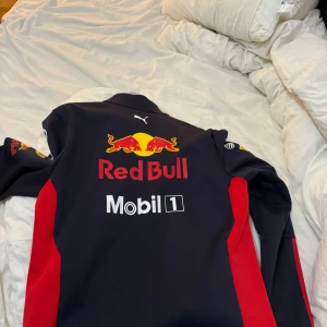 Svart och röd jacka från Puma - Säljer en cool svart och röd jacka från Puma med Red Bull-logga. Jackan har dragkedja och långa ärmar, perfekt för racingfans. Den har flera sponsorloggor som Aston Martin, Mobil 1 och Honda. Grym för dig som gillar motorsport!