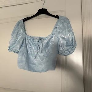 Ljusblå satinblus från NA-KD - Charmig ljusblå blus i satin från NA-KD med puffiga korta ärmar och en knytning framtill samt blommönster