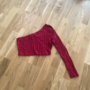 Röd oneshoulder topp - Snygg röd oneshoulder topp med ribbad textur. Perfekt för en trendig look. Den har en lång ärm och är croppad för en modern stil.