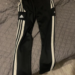 Svarta träningsbyxor från Adidas - Snygga svarta träningsbyxor från Adidas med vita ränder längs sidorna. Byxorna har en normal passform och är perfekta för sport och träning. De har en elastisk midja för extra komfort.