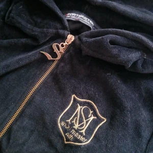 Svart hoodie från Sportmaster - Säljer en svart hoodie från Sportmaster med dragkedja och broderad logga på bröstet. Den har en huva och långa ärmar, perfekt för en casual look. Dragkedjan har en snygg detalj i form av en metallbokstav✨️