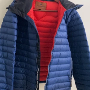 Blå pufferjacka från Jackson Hole - Säljer en blå pufferjacka från Jackson Hole med röd insida. Jackan har en dragkedja framtill och en huva för extra skydd. Perfekt för kyliga dagar! Pris kan diskuteras där vi både är glada
