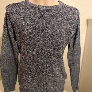 Melerad tröja från Clay Cooper - Snygg melerad tröja från Clay Cooper i en mix av blått och vit. Tröjan har långa ärmar och en rund halsringning med en dekorativ söm framtill. Perfekt för en stilren och avslappnad look.