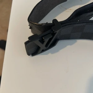 Svart rutigt bälte med monogramspänne - Snyggt svart bälte med ett diskret rutigt mönster och ett stilrent monogramspänne. Perfekt för att ge en touch av elegans till din outfit. Bältet har en modern design och passar till många olika stilar.