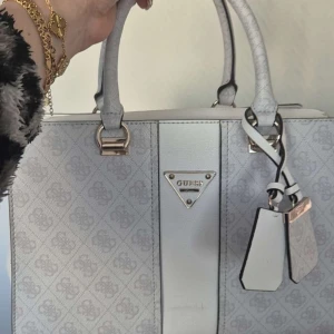 Vit handväska från Guess - Snygg vit handväska från Guess med ett diskret mönster och silverdetaljer. Väskan har dubbla handtag och en dekorativ rem med Guess-logga. Perfekt för att ge din outfit en stilren touch.
