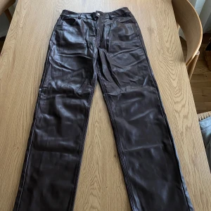 Bruna skinnbyxor från Gina Tricot - Snygga bruna skinnbyxor från Gina Tricot i storlek 38. Byxorna har en rak passform och är tillverkade i ett glansigt skinnmaterial. De har fickor både fram och bak samt en knapp och dragkedja i midjan.