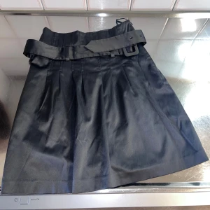 GråSvart kjol med bälte från Gina Tricot - Snygg gråsvart kjol från Gina Tricot med ett matchande bälte som ger en stilren look. Kjolen har en klassisk design med veckade detaljer och en kort längd. Perfekt för att skapa en chic outfit.