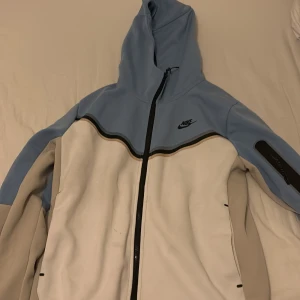 Blå och vit hoodie från Nike - Säljer en snygg hoodie från Nike i blått och vitt med dragkedja och huva. Jackan har en ficka med dragkedja på ärmen och Nike-loggan på bröstet. Perfekt för en sportig look!