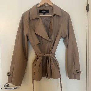 Beige trenchcoat från Vero moda - Snygg beige trenchcoat från Vero moda med knappar och knytskärp i midjan. Jackan har långa ärmar och en klassisk krage. Perfekt för en stilren look. Använt endast ett fåtal gånger och inköpt hösten 2024.