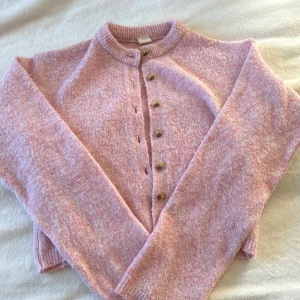 Rosa stickad kofta med knappar - Mysig rosa stickad kofta med rund hals och knappar framtill. Perfekt för kyliga dagar och ger en söt touch till din outfit. Koftan har långa ärmar och ribbade kanter.