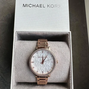 Roséguldfärgad klocka från Michael Kors - Snygg roséguldfärgad klocka från Michael Kors med glittriga detaljer runt urtavlan. Urtavlan har ett mönster med MK-loggan och är prydd med små stenar vid varje timme. Armbandet är i metall och justerbart med extra länkar. Perfekt för att ge en touch av lyx till din stil.
