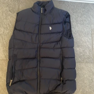 Mörkblå dunväst från U.S. Polo Assn. - Säljer en snygg mörkblå dunväst från U.S. Polo Assn. med dragkedja och broderad logga på bröstet. Perfekt för kyligare dagar och ger en stilren look.